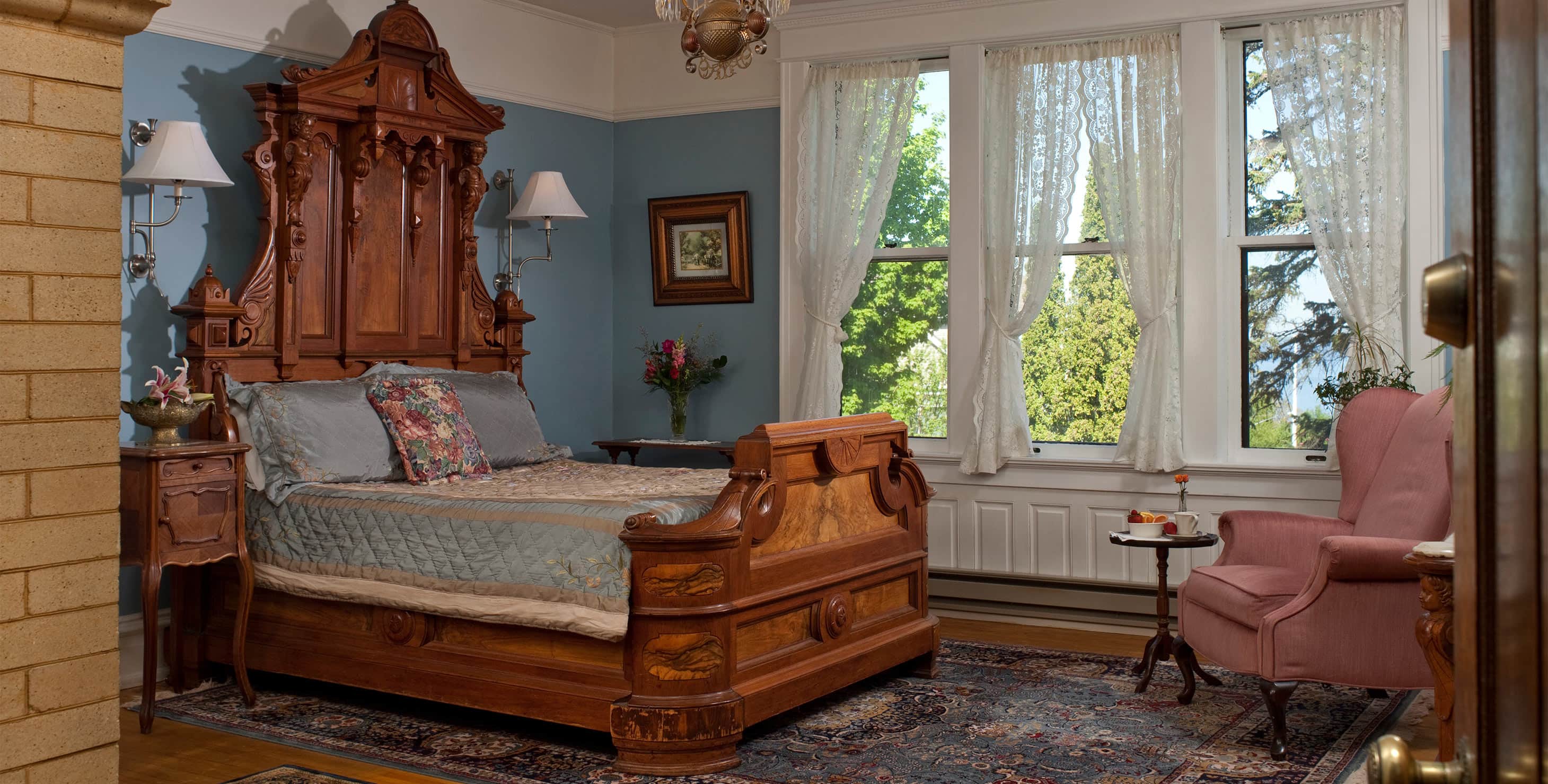 Ornate wood bed with side table, pink chair, and three windows