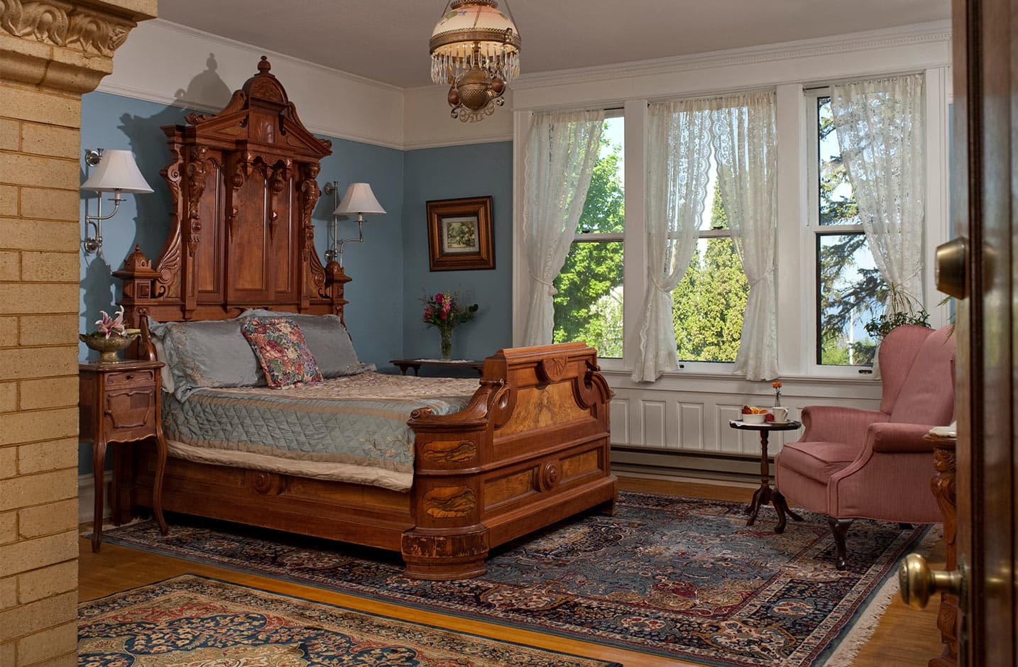 Ornate wood bed with side table, pink chair, and three windows
